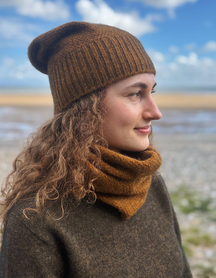 A woman outdoors wearing a brown ribbed knit beanie and a matching brown cowl neck warmer, with a blurred beach and blue sky in the background.