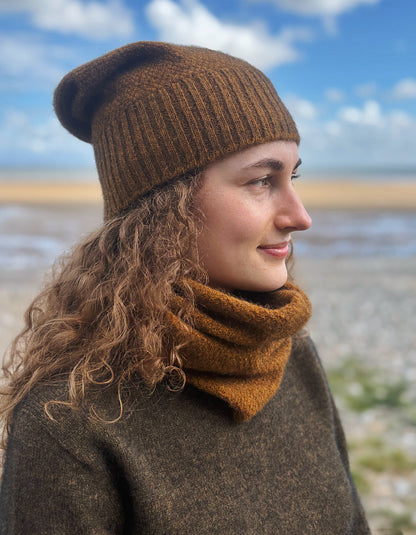 A woman outdoors wearing a brown ribbed knit beanie and a matching brown cowl neck warmer, with a blurred beach and blue sky in the background.
