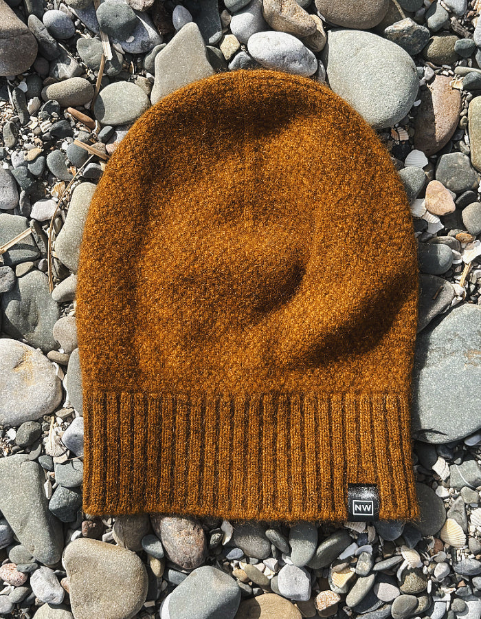 A textured knit beanie in a warm burnt orange-brown color, laid flat on a bed of smooth gray river stones. A small black brand tag is visible on the lower right edge of the beanie.