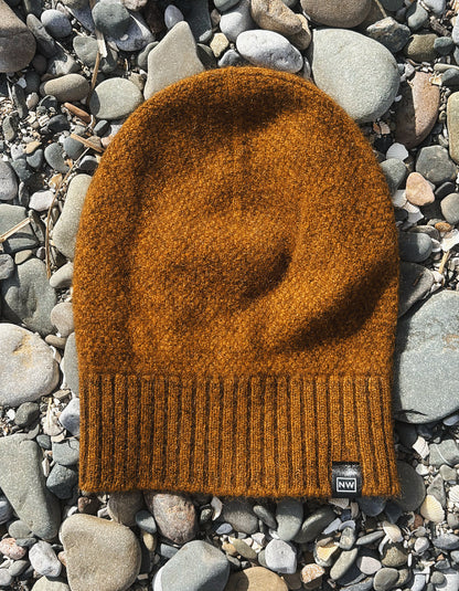 A textured knit beanie in a warm burnt orange-brown color, laid flat on a bed of smooth gray river stones. A small black brand tag is visible on the lower right edge of the beanie.