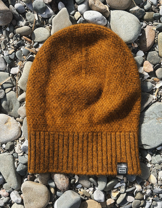 A textured knit beanie in a warm burnt orange-brown color, laid flat on a bed of smooth gray river stones. A small black brand tag is visible on the lower right edge of the beanie.