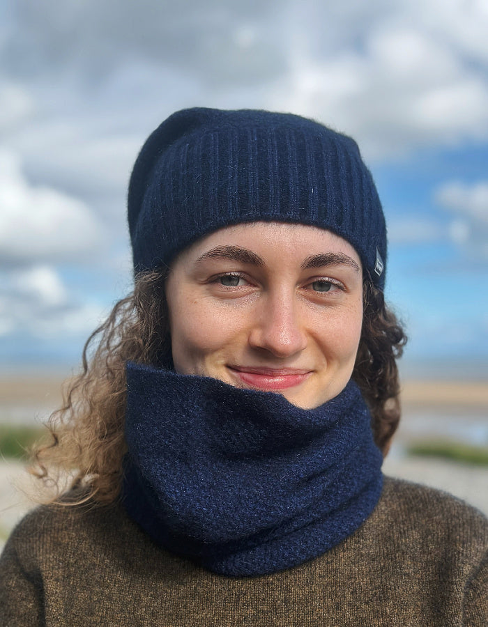 A woman outdoors wearing a dark navy blue ribbed knit beanie and matching navy blue neck gaiter, with a blurred sky and landscape background.