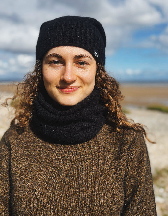 A woman outdoors wearing a black ribbed knit beanie with a small white logo tab and a matching black knit neck warmer, over a brown sweater, with a blurred beach and sky background.