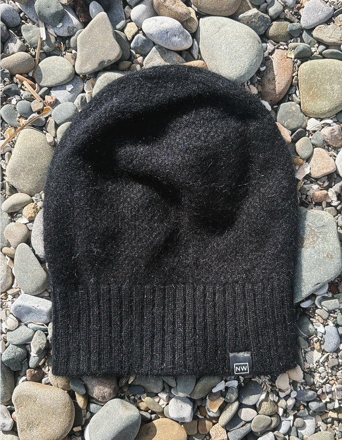 A black knit beanie hat with a ribbed cuff and a small square NW logo patch, laid flat on a bed of gray and tan river stones.