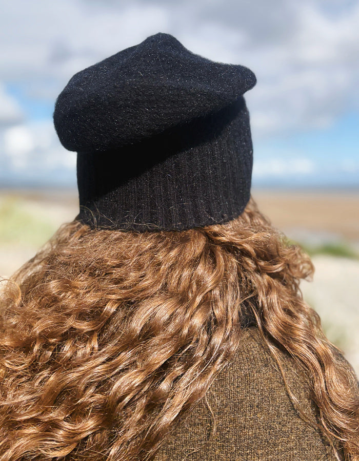 A person viewed from behind wearing a black knit beret and a brown textured coat, with long wavy light brown hair and an outdoor beach background.