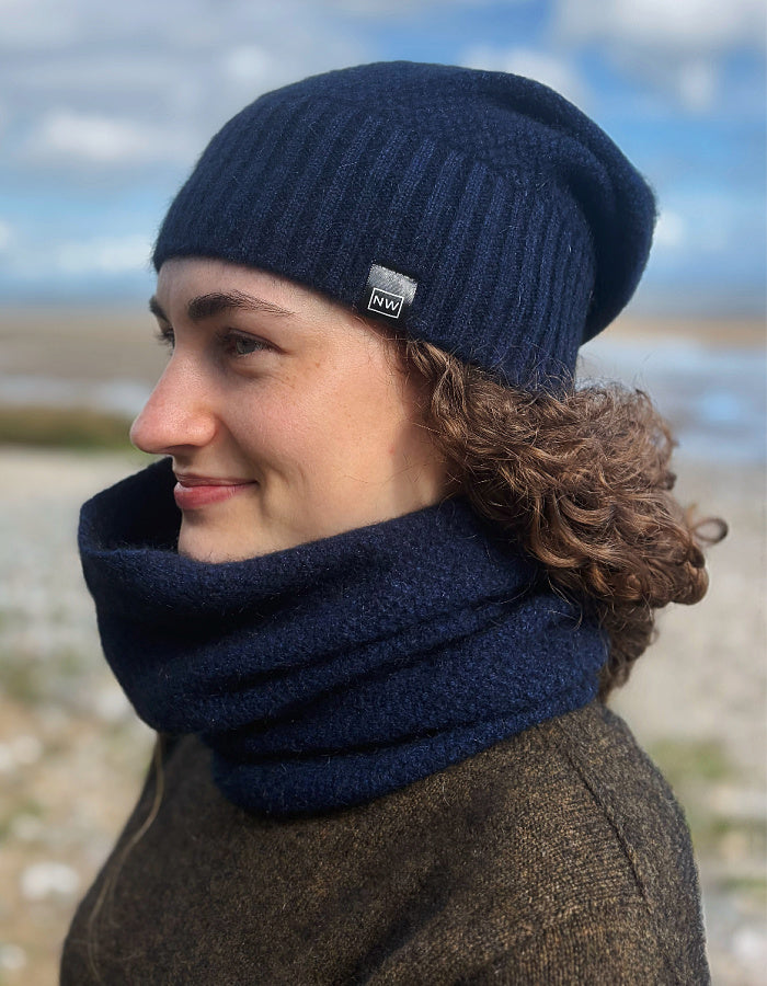 A person outdoors wearing a dark navy ribbed knit beanie with a small square brand patch and a matching navy cowl neck warmer.