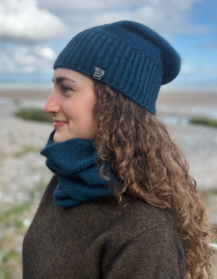 A woman in profile wearing a dark teal ribbed knit beanie and matching knit neck warmer outdoors on a rocky beach with cloudy sky in the background.
