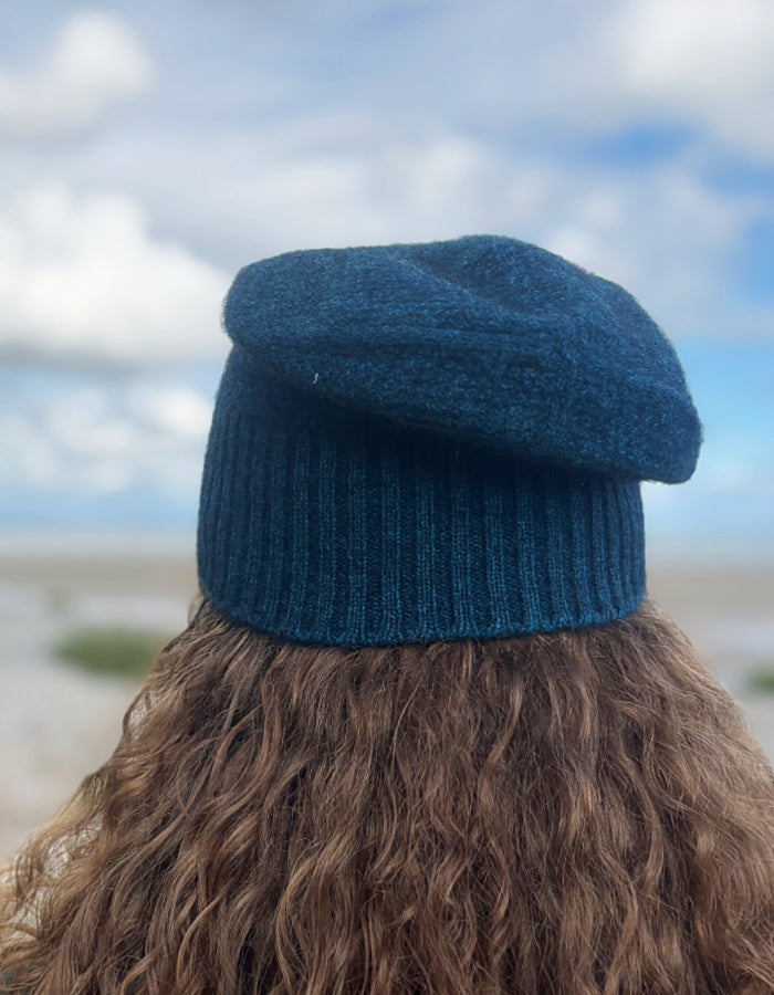 A person viewed from behind wearing a dark teal blue knit beret hat, with long brown curly hair and a blurred outdoor background of sky and sand.