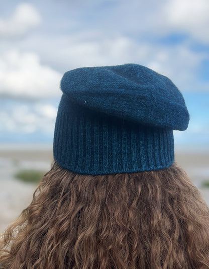 A person viewed from behind wearing a dark teal blue knit beret hat, with long brown curly hair and a blurred outdoor background of sky and sand.