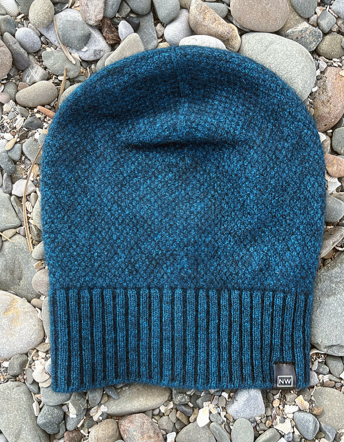 A textured knit beanie in a deep teal blue, laid flat on a bed of gray and white river stones. The beanie features a ribbed knit cuff and a small black square logo patch on the lower right side with the letters 'NW'.