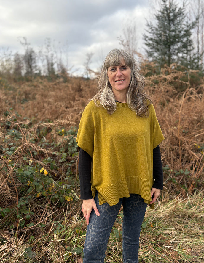 Woman wearing a mustard yellow poncho standing in a field with trees in the background