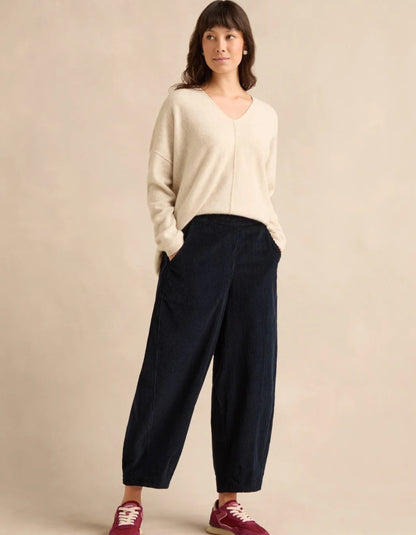 A woman modeling a cream knit V-neck sweater and dark navy wide-leg cropped pants, paired with burgundy sneakers, against a plain beige studio background.