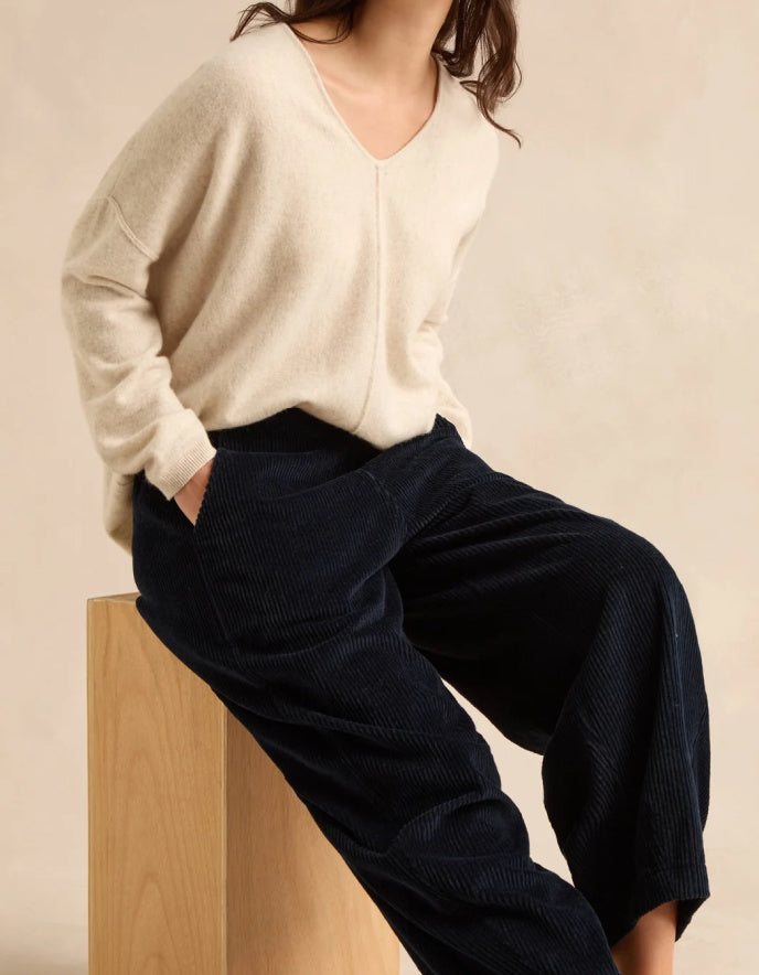 A model wearing a cream V-neck knit sweater and dark navy corduroy wide-leg pants, sitting on a light wood block against a warm beige background.