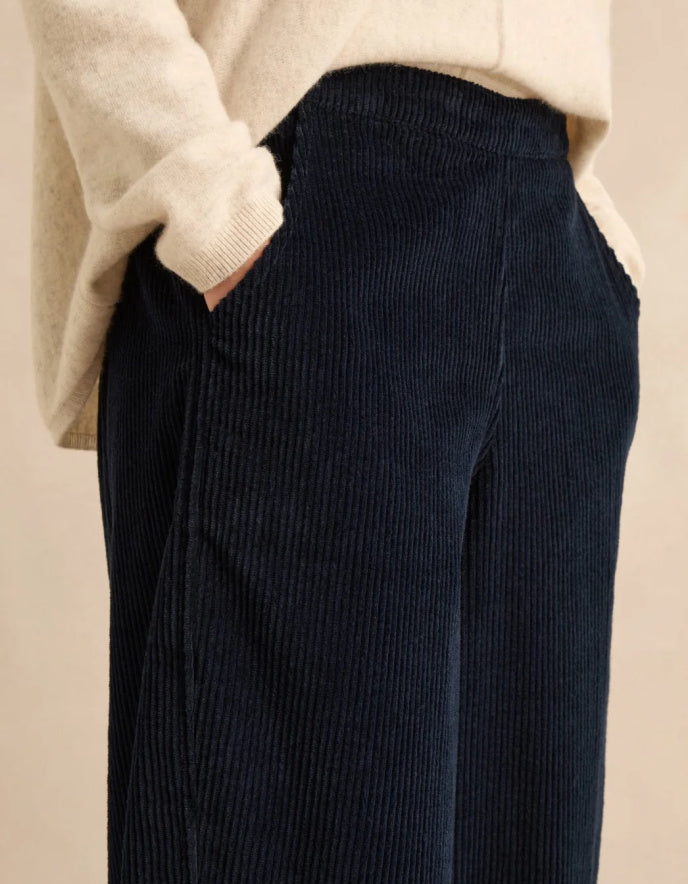 Close-up of a person wearing dark navy corduroy wide-leg trousers and a cream knit sweater, with one hand in a pocket, against a plain beige background.