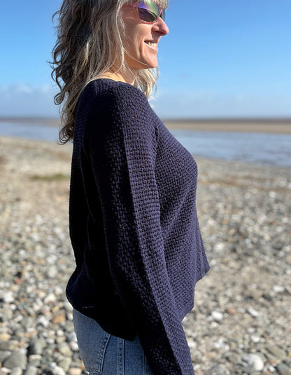 Woman wearing a dark blue knitted sweater on a pebbly beach with a clear sky.