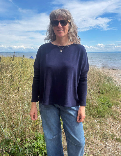 A woman with gray hair and sunglasses stands outdoors on a grassy shoreline with the sea and blue sky behind her. She is wearing a dark navy long-sleeve top and light blue jeans.