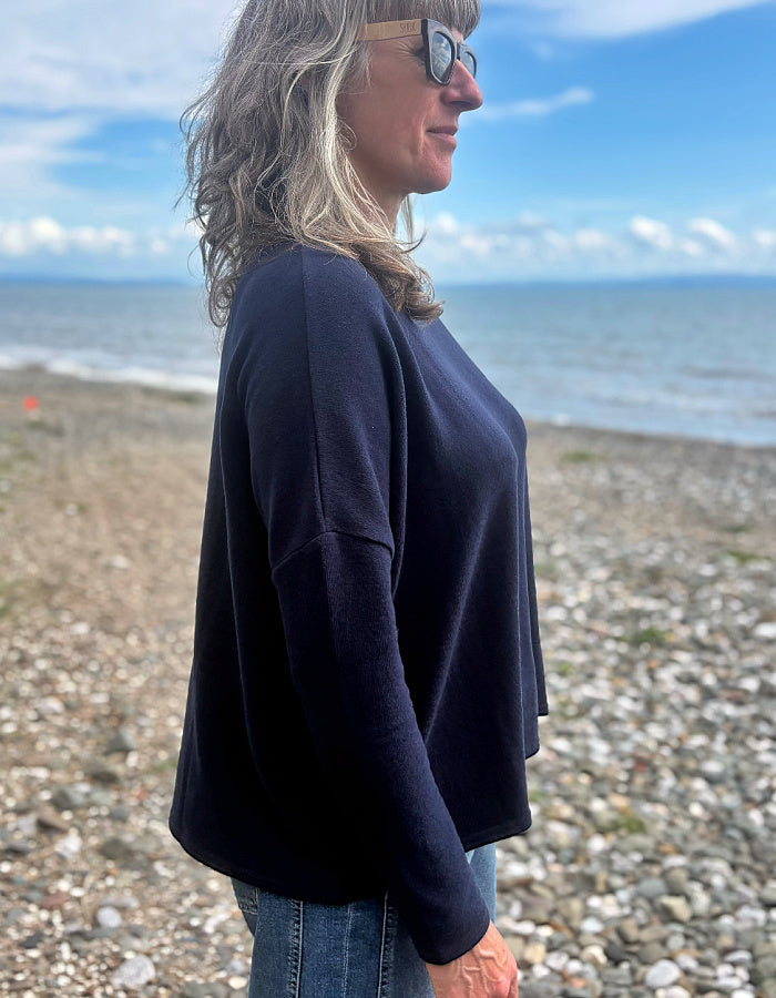 A woman with blonde-highlighted hair and sunglasses stands in profile on a pebble beach, wearing a loose, dark navy long-sleeve top and blue jeans, with the ocean and blue sky in the background.