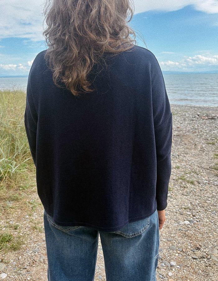 A person viewed from behind wearing a dark navy blue long-sleeve top and blue jeans, standing on a pebble beach with grass and water in the background.