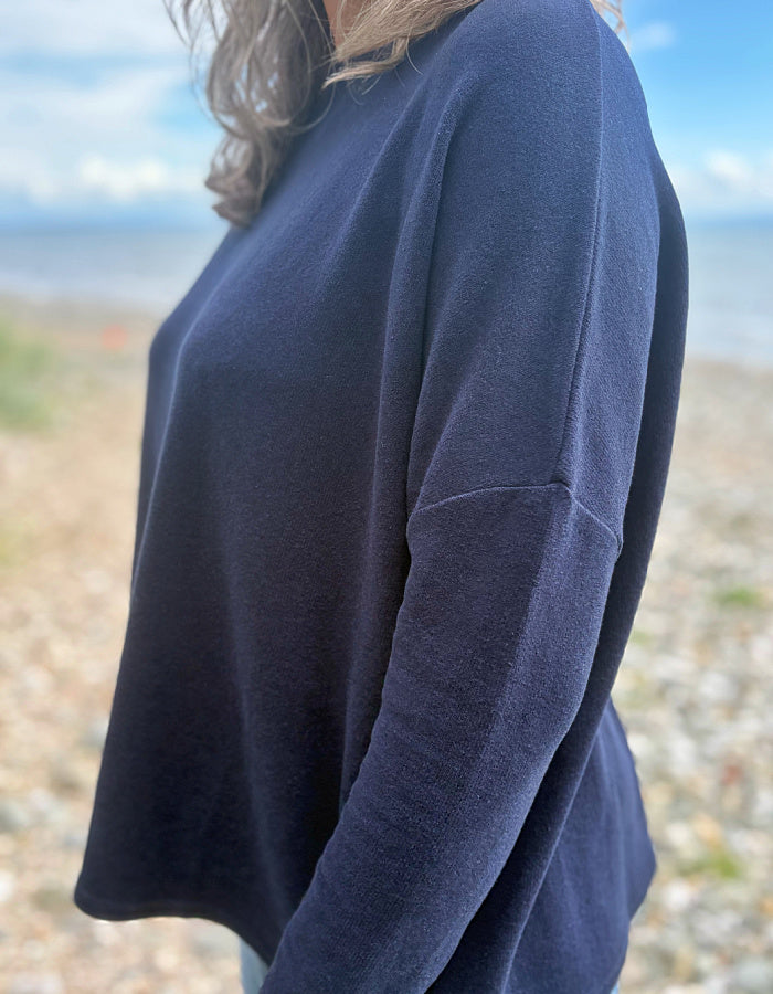 A person wearing a loose, long-sleeve navy knit top standing outdoors on a pebble beach with water and sky blurred in the background.