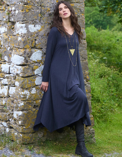 Woman in a long dark blue dress standing against a stone wall with greenery in the background