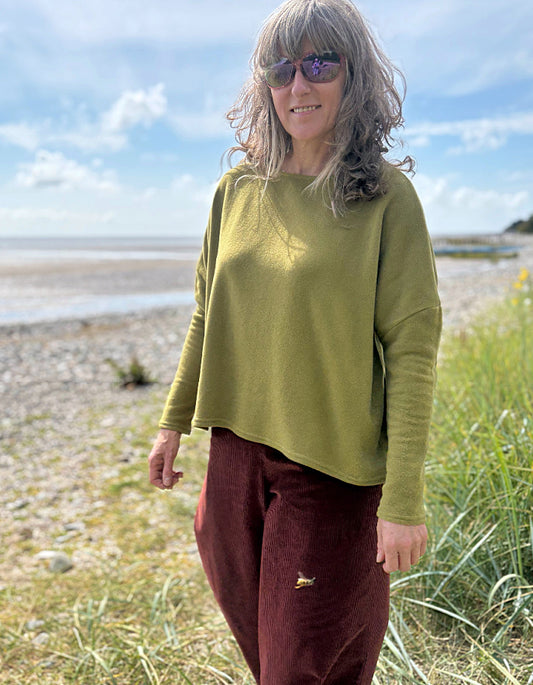 Person wearing a green sweater and maroon pants standing on a beach with grass and water in the background.