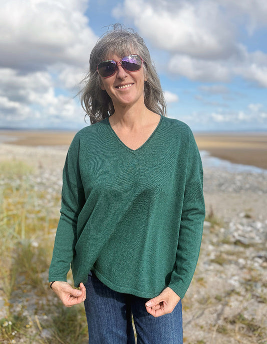 Person wearing a green V neck merino sweater standing in a scenic outdoor setting with a blue sky.