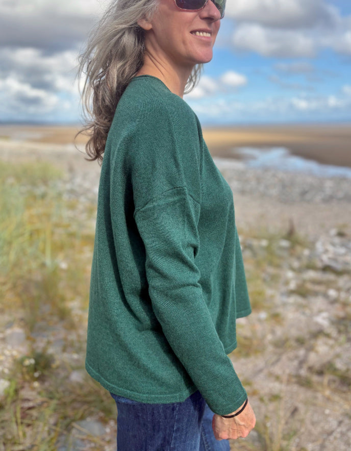 Person wearing a green V neck merino sweater standing in a scenic outdoor setting with a blue sky.