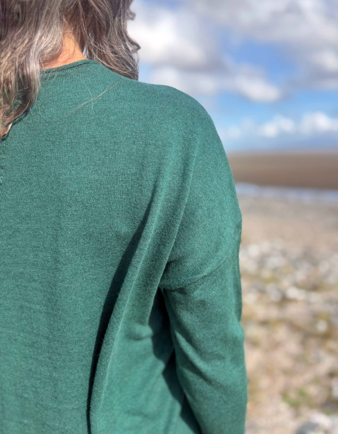 Person wearing a green V neck merino sweater standing in a scenic outdoor setting with a blue sky.