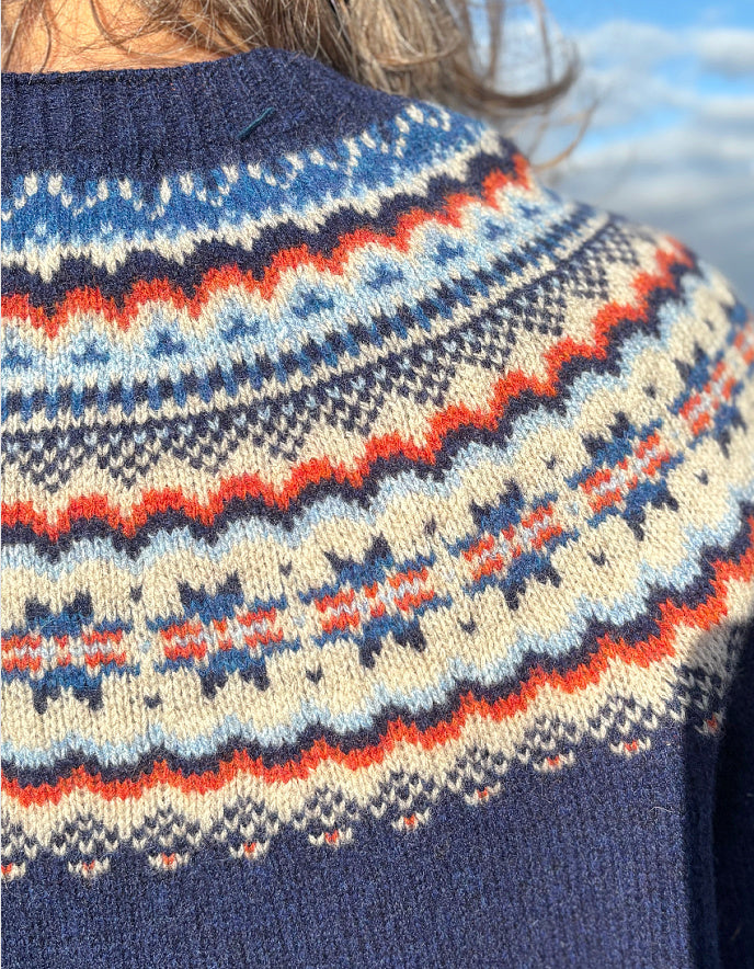 Close-up of a person wearing a patterned sweater with a blue, red, and white design against a blurred natural background.