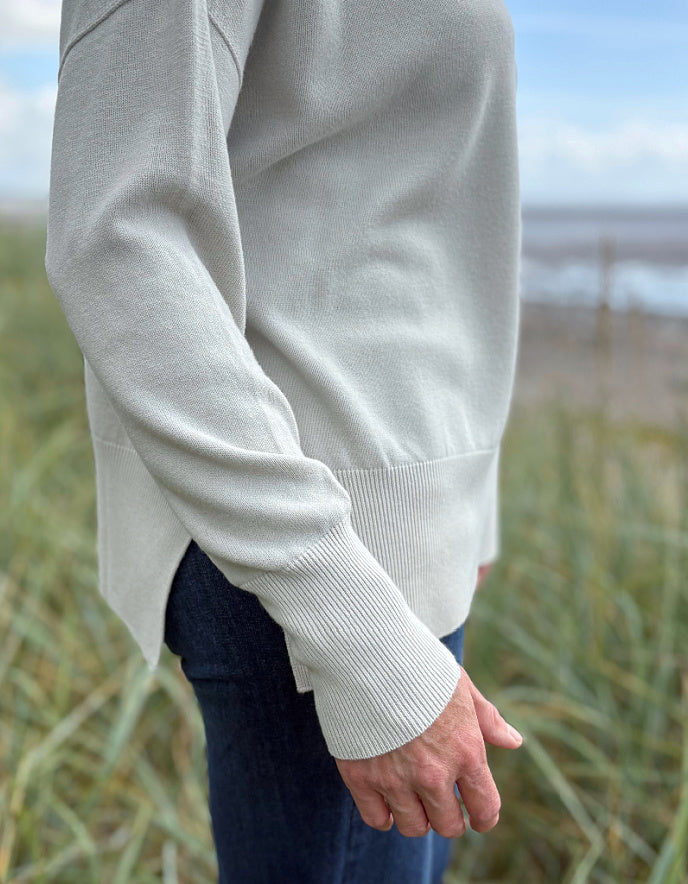 Woman standing in beach grass wearing a light-coloured cream turtle neck sweater and sunglasses with a beach and sky in the background.