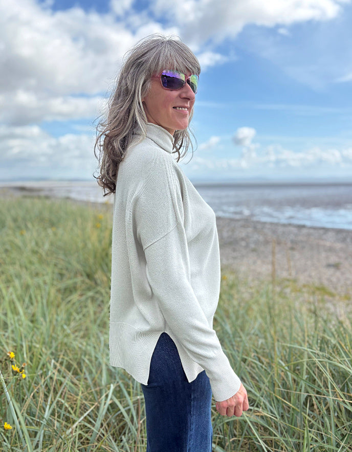 Woman standing in beach grass wearing a light-coloured cream turtle neck sweater and sunglasses with a beach and sky in the background.