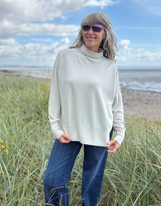 Person standing in grass near a beach wearing a light-colored sweater and sunglasses.