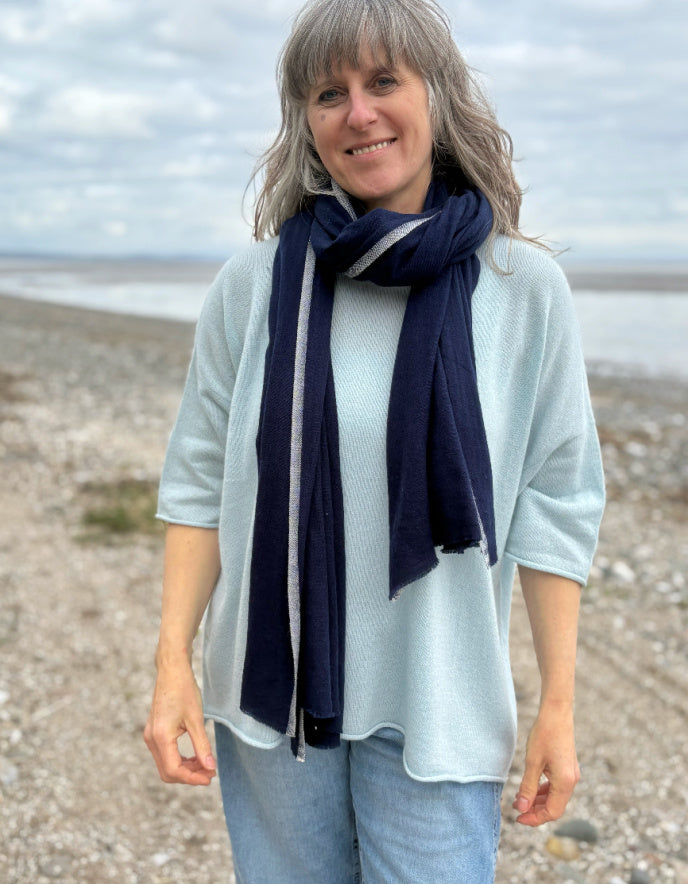 Woman wearing a light blue sweater and navy scarf standing on a beach.