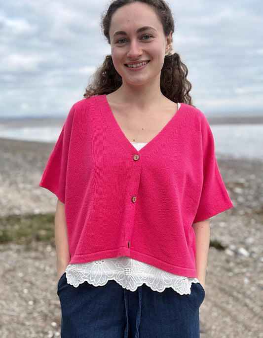 Woman wearing a bright pink cardigan over a white lace top on a beach.