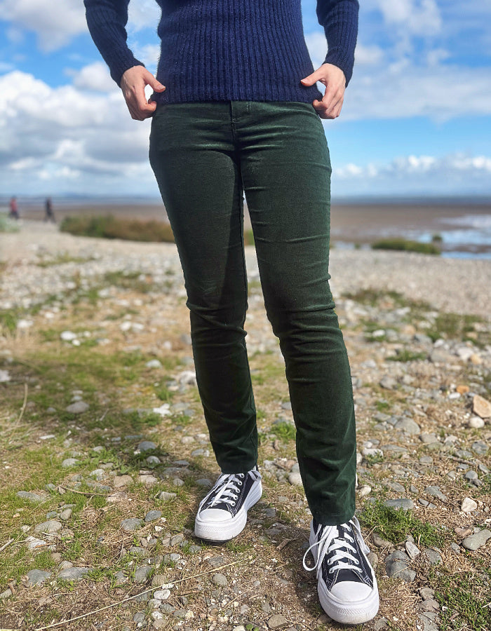 A person standing outdoors on a rocky shoreline wearing dark green slim-fit pants, a navy ribbed sweater, and black-and-white sneakers.