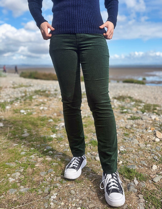 A person standing outdoors on a rocky shoreline wearing dark green slim-fit pants, a navy ribbed sweater, and black-and-white sneakers.