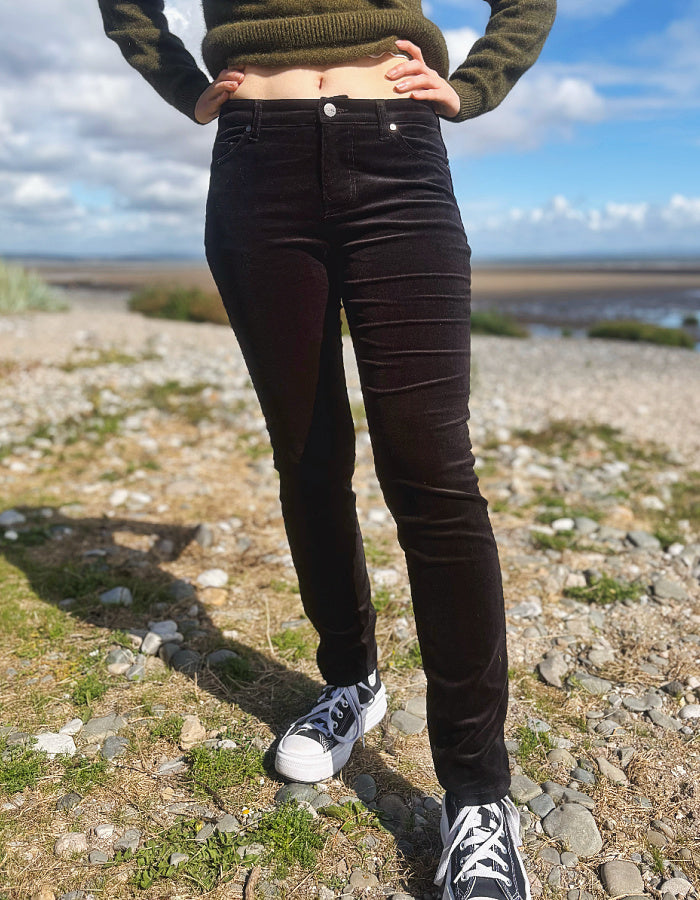 A person outdoors wearing dark brown corduroy pants, a dark olive green knit sweater, and black-and-white sneakers, standing on a rocky, grassy shoreline under a blue sky with clouds.