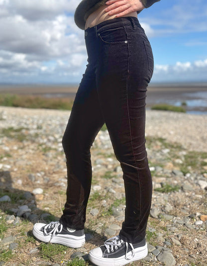 A person outdoors wearing slim-fit black corduroy jeans and black-and-white platform sneakers, standing on a rocky shoreline with water and cloudy sky in the background.
