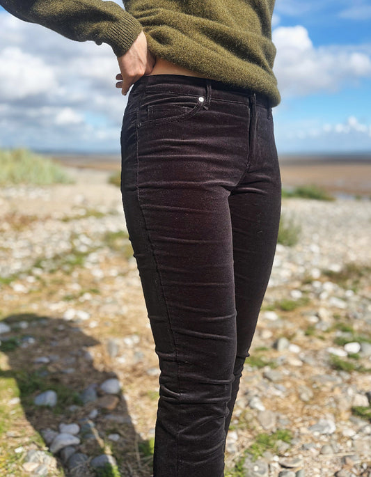 A person outdoors wearing dark brown corduroy skinny jeans and an olive green sweater, standing on a rocky beach with a cloudy blue sky in the background.