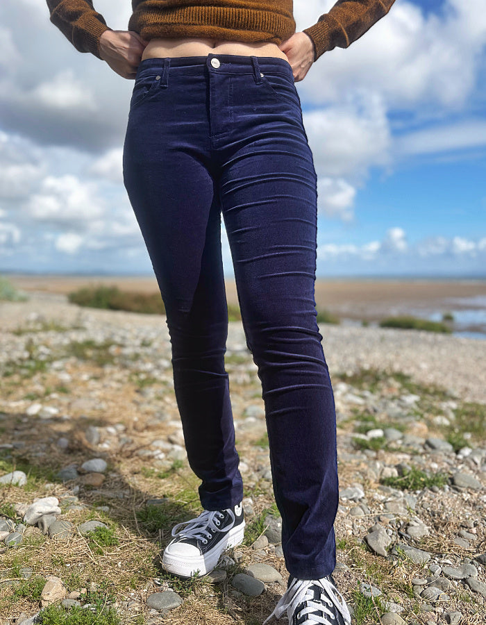 A person outdoors wearing dark navy blue skinny jeans, a brown knit sweater, and white sneakers, standing on a rocky shoreline under a cloudy blue sky.