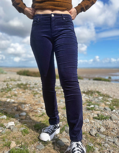 A person outdoors wearing dark navy blue skinny jeans, a brown knit sweater, and white sneakers, standing on a rocky shoreline under a cloudy blue sky.