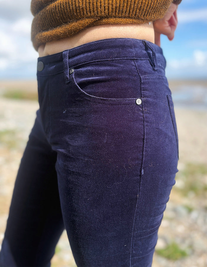 A close-up side view of a person wearing dark navy blue velvet or velour pants, paired with a brown knit sweater, standing outdoors on a sandy beach with water and sky in the background.