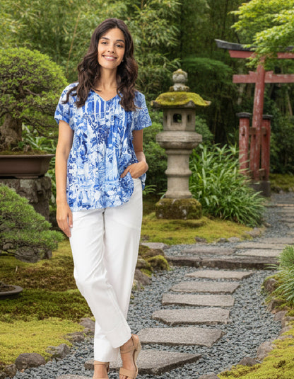 Woman in a blue floral blouse and white pants standing in a garden with stone lanterns and a torii gate.