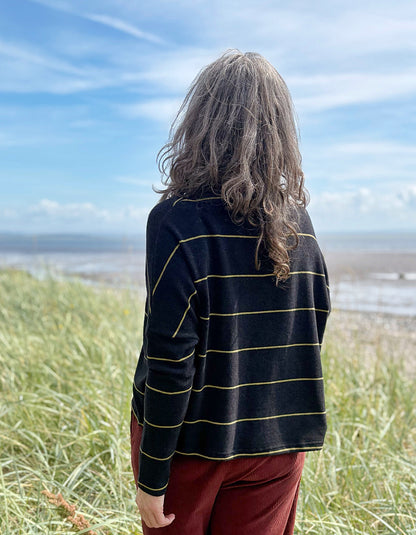 Person wearing a black and yellow striped shirt standing on grassy dunes with a beach and blue sky in the background
