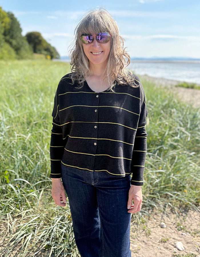 Woman wearing charcoal and olive pin stripe cardigan and sunglasses, standing in a grassy field with a body of water in the background.
