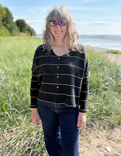 Woman wearing charcoal and olive pin stripe cardigan and sunglasses, standing in a grassy field with a body of water in the background.
