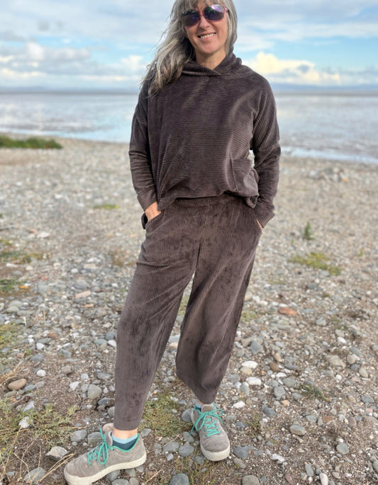 Person wearing a brown cord lantern shaped trousers standing on a pebbly beach with ocean in the background