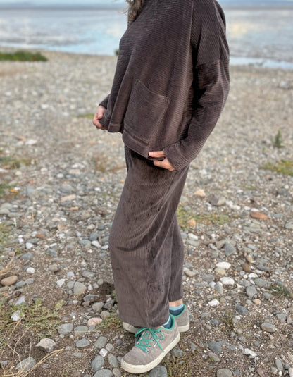 Person wearing a brown cord lantern shaped trousers standing on a pebbly beach with ocean in the background