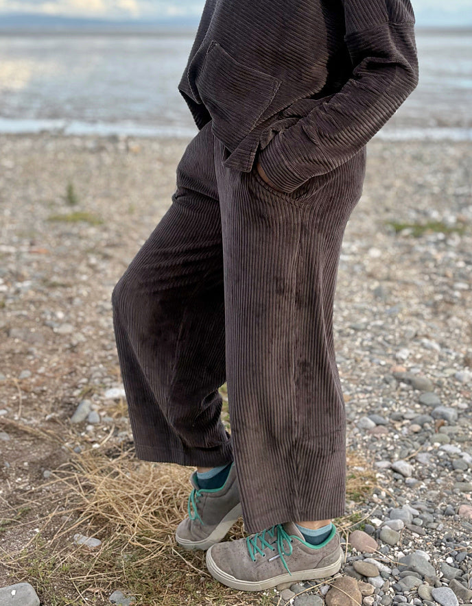 Person wearing a brown cord lantern shaped trousers standing on a pebbly beach with ocean in the background