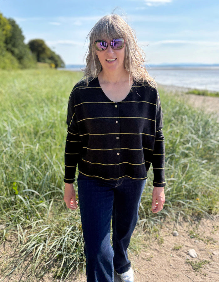 Woman wearing charcoal and olive pin stripe cardigan and sunglasses, standing in a grassy field with a body of water in the background.
